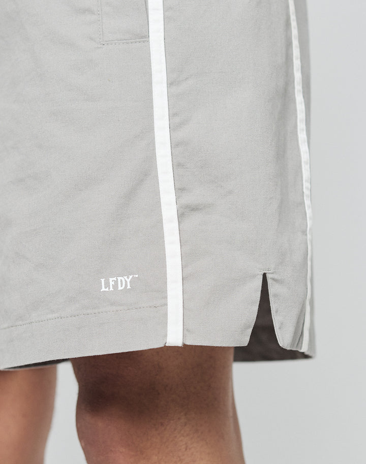Close-up of a person wearing LIVE FAST DIE YOUNG Basic Cotton Shorts in light gray with a white vertical stripe, elastic waistband, LFDY logo print near the hem, small side slit, and a comfortable regular fit.
