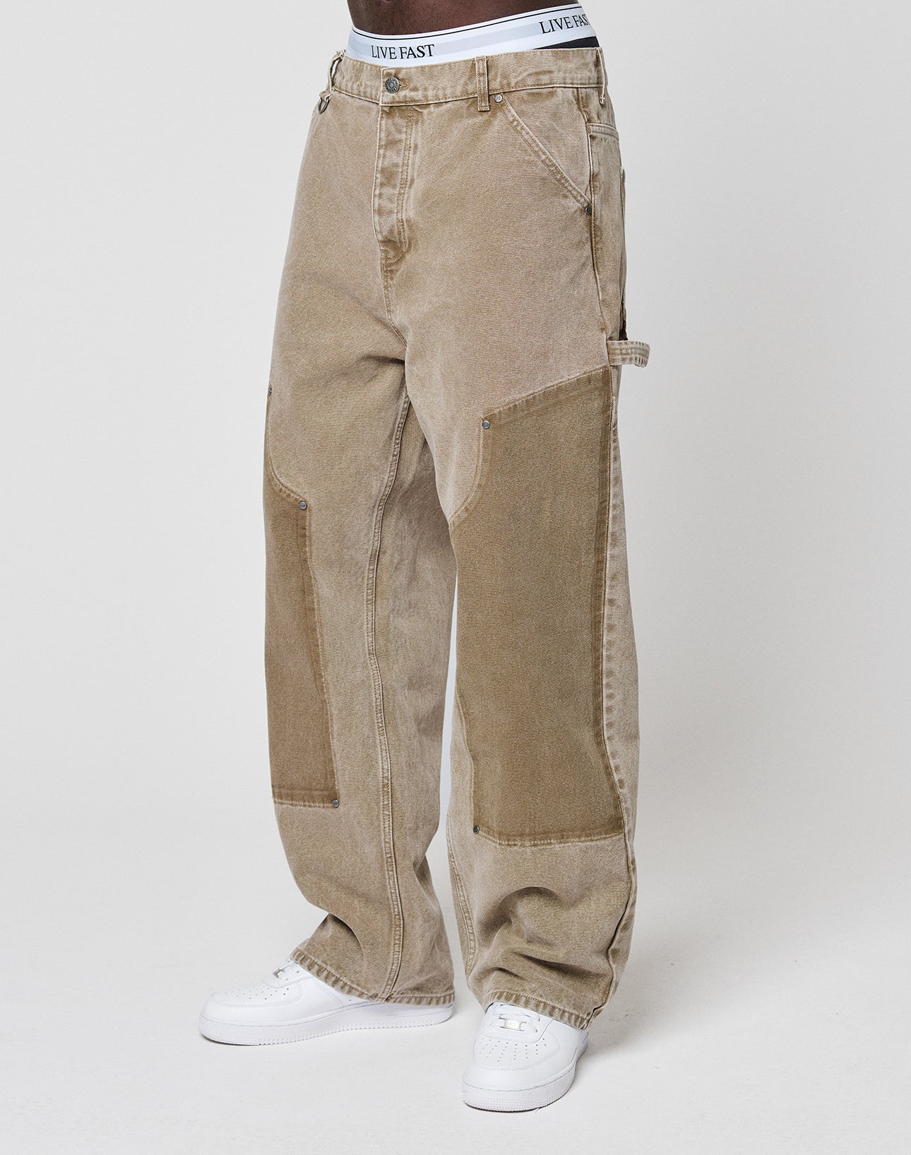 A person in LIVE FAST DIE YOUNG Faded Carpenter Pants with vintage wash and reinforced knees, white LIVE FAST waistband visible, and white sneakers, stands against a plain light background.
