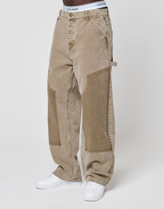 A person in LIVE FAST DIE YOUNG Faded Carpenter Pants with vintage wash and reinforced knees, white LIVE FAST waistband visible, and white sneakers, stands against a plain light background.