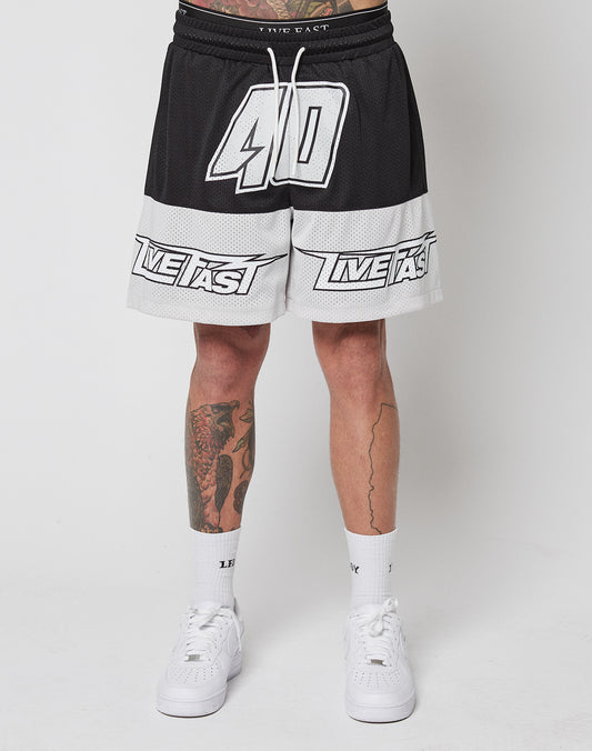 A person wears LFDY Racing Mesh Shorts by LIVE FAST DIE YOUNG, featuring a black-and-white design with “40” and “LIVE FAST” prints, a Regular Fit, and elastic waistband. Paired with white socks, white sneakers, and visible leg tattoos.