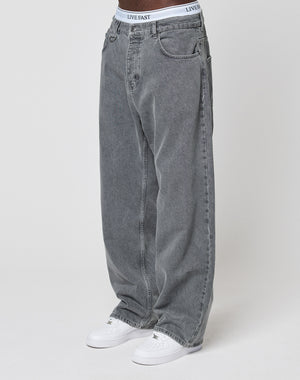 A person wearing LFDY Baggy Denim jeans by LIVE FAST DIE YOUNG, styled with white sneakers and visible white underwear featuring a LIVE FAST waistband, stands against a plain white background.