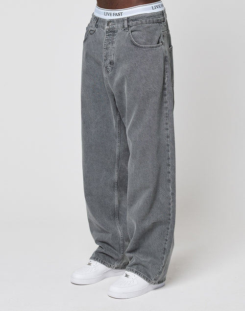 A person wearing LFDY Baggy Denim jeans by LIVE FAST DIE YOUNG, styled with white sneakers and visible white underwear featuring a LIVE FAST waistband, stands against a plain white background.