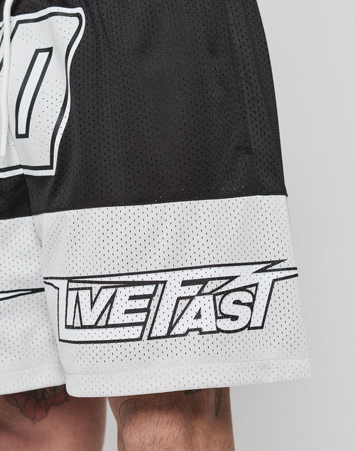 Close-up of a person wearing black and white LFDY Racing Mesh Shorts by LIVE FAST DIE YOUNG, featuring bold lettering across the lower leg. The regular fit polyester shorts have an elastic waistband and reveal part of a tattooed leg.