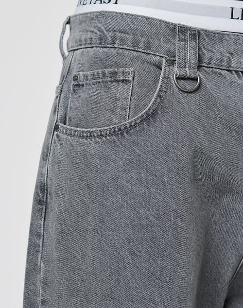 Close-up of a person wearing LFDY Baggy Denim jeans by LIVE FAST DIE YOUNG in light gray with baggy fit, straight leg, small front pocket, belt loops, silver D-ring, and white underwear waistband with black text peeking above.