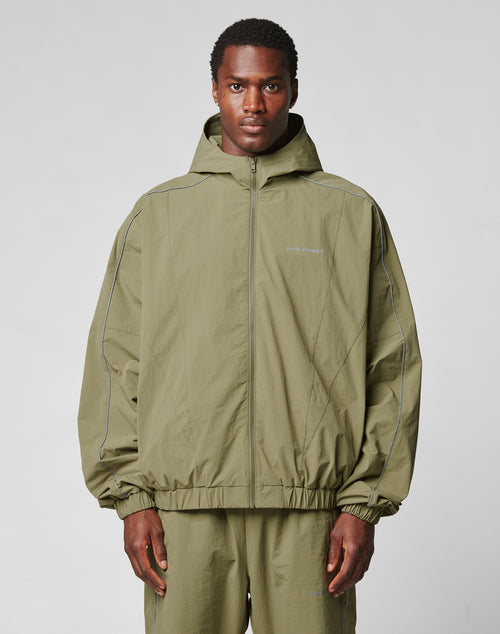 A man wears the LIVE FAST DIE YOUNG Live Fast Trackjacket in olive green nylon with reflective piping, a front zipper, and matching pants, standing against a plain light gray background.