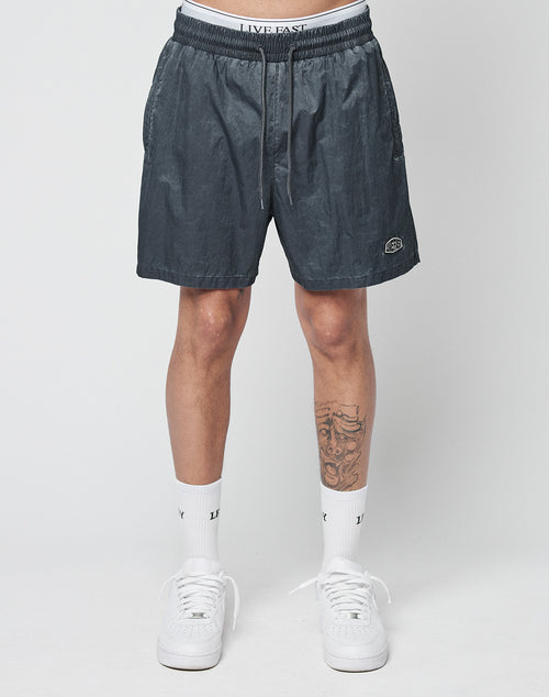 A person stands against a plain background wearing LIVE FAST DIE YOUNG Live Fast Swimshorts in charcoal gray, white LIVE FAST elastic waistband underwear, white socks with LV text, and white sneakers. A tattoo is visible on their left shin.