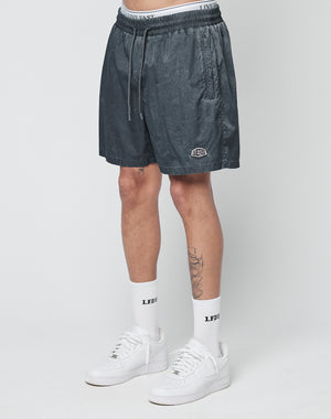 A person wears LIVE FAST DIE YOUNG Live Fast Swimshorts in dark gray (regular fit, elastic waistband, logo patch on left leg) with white logo socks and sneakers, standing against a white background. Part of a leg tattoo is visible.