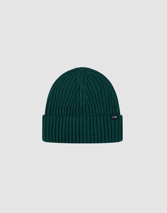 The LIVE FAST DIE YOUNG Rib Knit Beanie in dark green is made from soft polyacryl, with a folded cuff and small black side tag. Displayed on a plain light gray background. One size for versatile comfort.