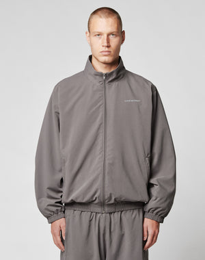 A young man with short hair wears the LIVE FAST DIE YOUNG LFDY Trackjacket and matching polyester pants in gray, standing against a plain light gray background and looking at the camera with a neutral expression.