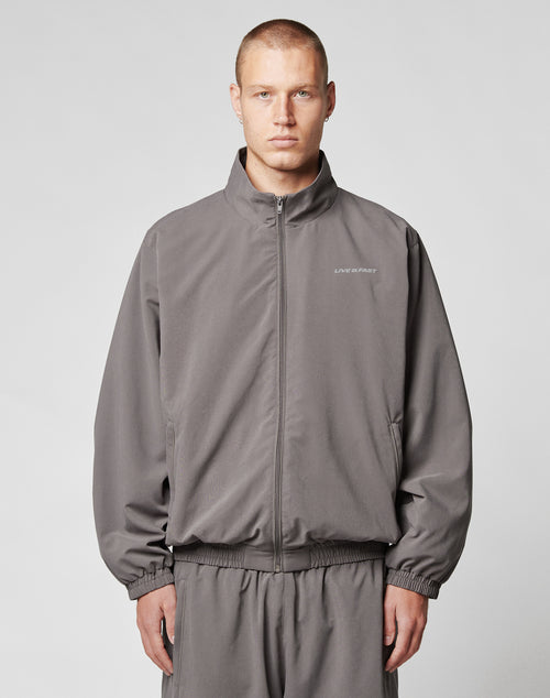 A young man with short hair wears the LIVE FAST DIE YOUNG LFDY Trackjacket and matching polyester pants in gray, standing against a plain light gray background and looking at the camera with a neutral expression.