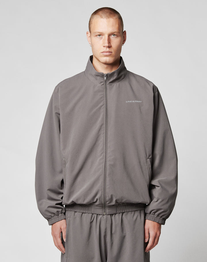 A young man with short hair wears the LIVE FAST DIE YOUNG LFDY Trackjacket and matching polyester pants in gray, standing against a plain light gray background and looking at the camera with a neutral expression.