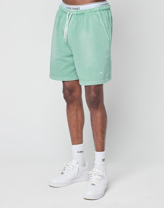 A person models light green Basic Sweat Shorts by LIVE FAST DIE YOUNG, featuring a regular fit, drawstring waist, and LIVE text near the hem. Only the lower half is shown, styled with white socks and sneakers against a plain white background.