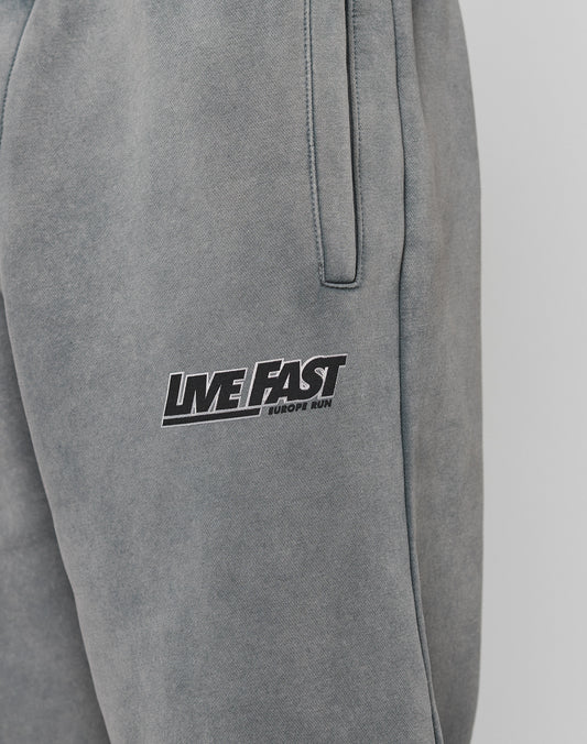 Close-up of LFDY Europe Run Jogger by LIVE FAST DIE YOUNG. Grey, loose fit sweatpants with side pockets, elastic cuffs, and LIVE FAST EUROPE RUN printed in black and white on the upper thigh.