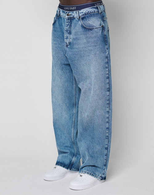A person wears the LFDY Baggy Denim by LIVE FAST DIE YOUNG—baggy, light blue jeans with a wide straight leg and baggy fit, made of 100% cotton—paired with a black LIVE FAST waistband and white sneakers against a white background.