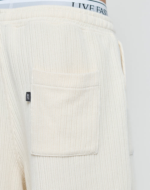 Close-up of cream-colored Basic Knit Shorts by LIVE FAST DIE YOUNG, featuring ribbed knit, patch pockets, and a small black tag. The elastic waistband of underwear with the text LIVE FAST is visible above the shorts.