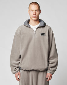 A person with short hair wears the LIVE FAST DIE YOUNG Live Fast Fleece Halfzip, a loose, light gray fleece half-zip pullover with an oversized fit and small left chest logo, standing against a plain light background. | model