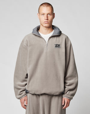 A person with short hair wears the LIVE FAST DIE YOUNG Live Fast Fleece Halfzip, a loose, light gray fleece half-zip pullover with an oversized fit and small left chest logo, standing against a plain light background.