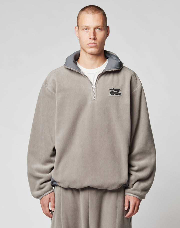 A person with short hair wears the LIVE FAST DIE YOUNG Live Fast Fleece Halfzip, a loose, light gray fleece half-zip pullover with an oversized fit and small left chest logo, standing against a plain light background. | model