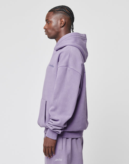A man with braided hair stands in profile, wearing the Basic 360 Hooded and matching sweatpants by LIVE FAST DIE YOUNG. This oversize fit lavender hoodie is made from Baumwolle Polyester, with a front pocket and subtle chest text on white.