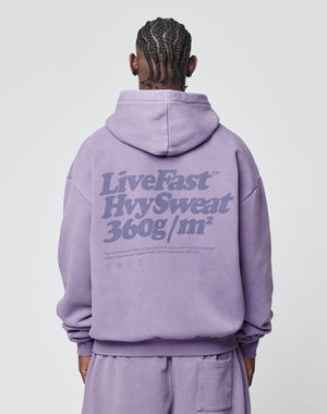Seen from behind, a person with braided hair wears the LIVE FAST DIE YOUNG Basic 360 Hooded in Oversize Fit, lavender, with bold back text LiveFast HvySweat 360g/m² and smaller print below. Vintage-washed Baumwolle-Polyester blend.
