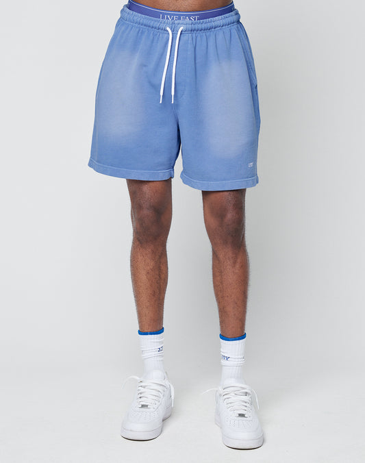 A person stands facing forward against a white background, wearing LIVE FAST DIE YOUNG Basic Sweat Shorts in light blue with a faded look, white drawstring and elastic waistband, paired with white socks and sneakers.