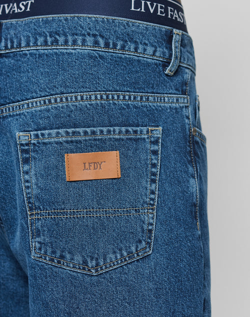 Close-up of Wide Denim jeans by LIVE FAST DIE YOUNG: blue denim with a brown LFDY patch, 100% cotton, regular fit with a stylish wide leg. Navy waistband shows LIVE FAST in white on the underwear.