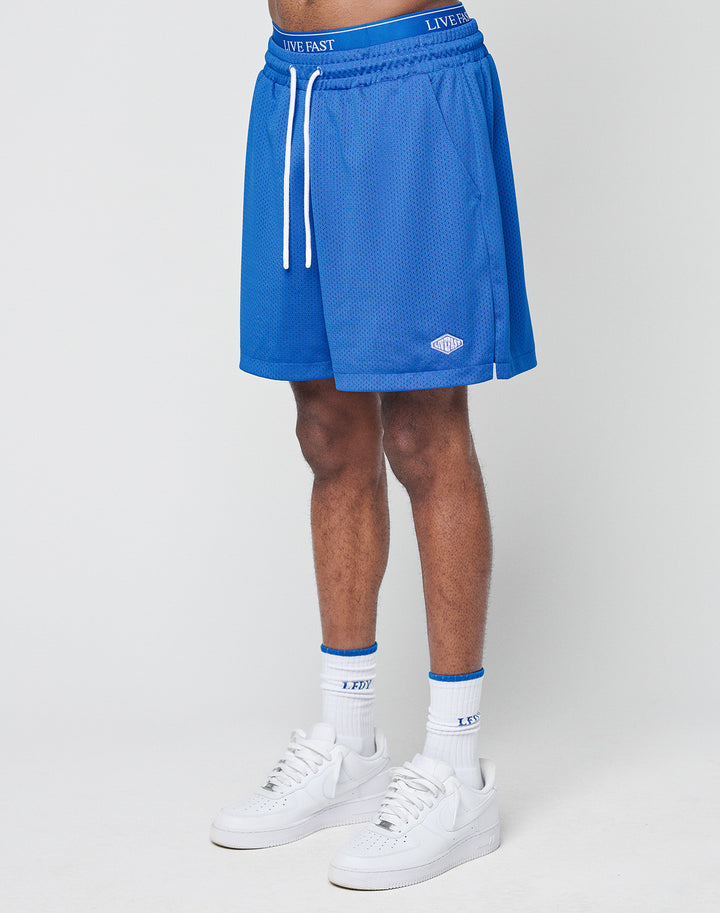 A person wears blue Basic Mesh Shorts by LIVE FAST DIE YOUNG with an elastic waistband and drawstring featuring LIVE FAST, paired with white socks with blue trim and white sneakers, standing against a plain white background.