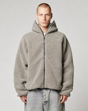 A person with short hair wears a light gray Live Fast Teddy Jacket by LIVE FAST DIE YOUNG, featuring subtle embossed branding and a high collar, paired with loose, faded jeans against a plain, light background.