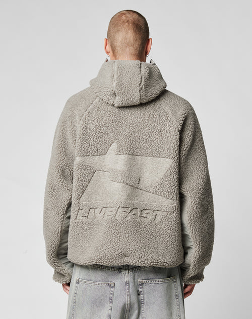 Seen from behind, a person with short hair wears the LIVE FAST DIE YOUNG Live Fast Teddy Jacket in gray fleece with a hood, featuring embossed branding and the word LIVEFAST on the back, paired with light gray jeans.