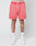 A person wears LIVE FAST DIE YOUNG Basic Sweat Shorts in coral pink with an elastic waistband and handcrafted fade effect, white socks with red accents, and white sneakers. Only their lower body is visible against a plain light background.