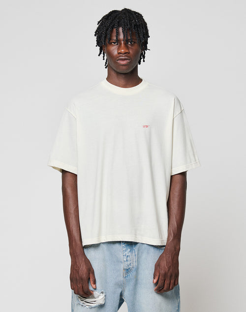 A young man with short twists models the LIVE FAST DIE YOUNG LFDY Flash Tee, a loose-fitting off-white 160gsm shirt with LFTY chest print, paired with light blue ripped jeans against a plain light gray background.