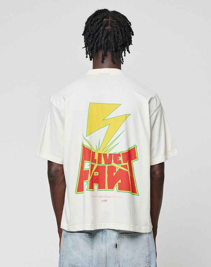 A person with short twists wears the LFDY Flash Tee by LIVE FAST DIE YOUNG—a white, 160gsm oversize T-shirt with a yellow lightning bolt and bold red LIVE FAST text on the back—paired with light blue jeans against a plain background. | model