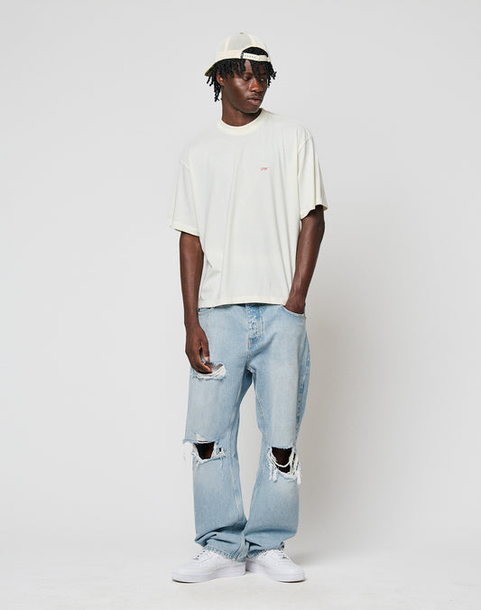 A man wearing a light beige 100% Baumwolle t-shirt, LFDY Distressed Baggy Denim by LIVE FAST DIE YOUNG, white sneakers, and a backwards white cap stands against a white background, looking to the side with one hand in his pocket.