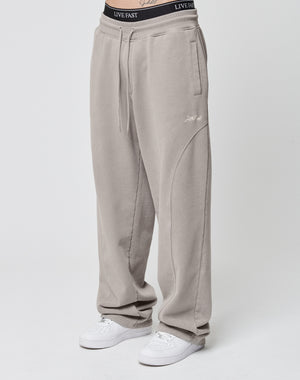 Someone wearing the LIVE FAST DIE YOUNG Basic Wide Jogger in light taupe, featuring a wide fit, subtle stitched details, and an elastic waistband. Paired with white sneakers and a visible black LIVE FAST waistband. Plain white background.