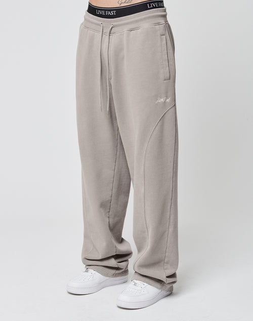 Someone wearing the LIVE FAST DIE YOUNG Basic Wide Jogger in light taupe, featuring a wide fit, subtle stitched details, and an elastic waistband. Paired with white sneakers and a visible black LIVE FAST waistband. Plain white background.