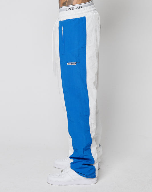 Side view of a person wearing LIVE FAST DIE YOUNG LFDY Trackpants 3000: wide fit, white with bold blue stripe, LIVE FAST on elastic waistband, multi-logo details, zip pocket, LVFST patch—styled with classic white sneakers.