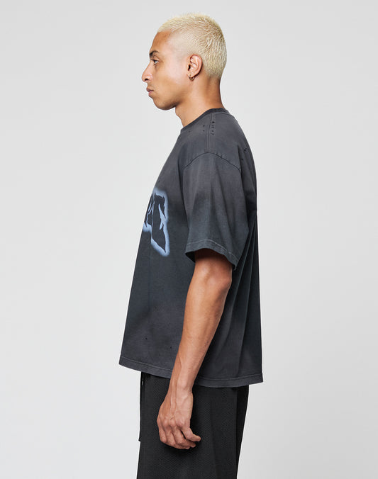 A man with short bleached blonde hair stands in profile against a plain light background, wearing the LFDY Flames Tee by LIVE FAST DIE YOUNG—an oversize fit black graphic T-shirt with handgefertigter Sprayeffekt—and black shorts.