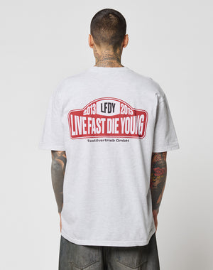 A tattooed person, seen from behind, wears the LIVE FAST DIE YOUNG Live Fast 2013 Tee in light gray with a bold red and black LFDY LIVE FAST DIE YOUNG 2013 graphic on the back.