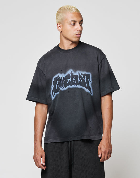 A person with short, bleached blond hair wears the LIVE FAST DIE YOUNG LFDY Flames Tee, a loose black oversized t-shirt with a bold spray effect graphic on the chest, paired with black shorts against a plain light background.