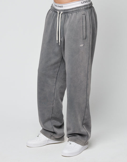 A person wears the LIVE FAST DIE YOUNG Basic Wide Jogger in Oversize Fitāloose gray sweatpants with white drawstrings and an elastic waistband showing LIVE FAST textāpaired with white sneakers against a plain light gray background.