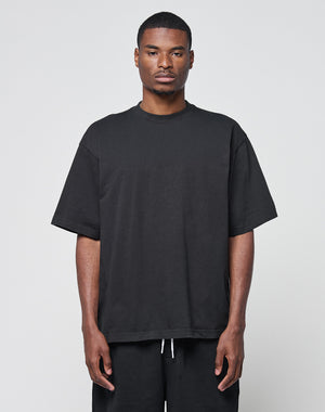 A man faces forward against a plain light gray background, wearing the LIVE FAST DIE YOUNG Basic Heavy Tee in oversize black 100% Baumwolle and black pants, with a neutral expression and arms relaxed at his sides.