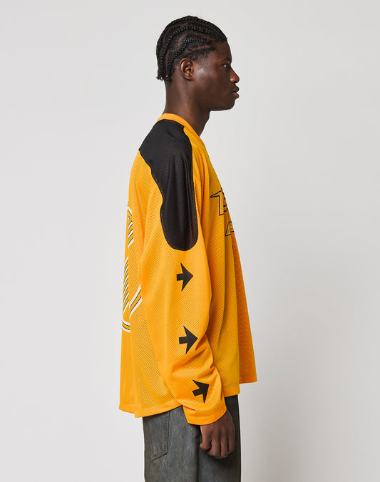 A person with braided hair stands in profile against a plain background, wearing the LIVE FAST DIE YOUNG LFDY Autobahn Jersey—an oversized yellow and black long-sleeve shirt with arrow graphics on the sleeves—and dark pants.