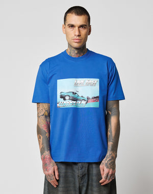 A tattooed man wears the LIVE FAST DIE YOUNG LFDY Crash Kid Tee, a blue regular fit T-shirt with a car graphic and text, standing facing forward against a plain light background.