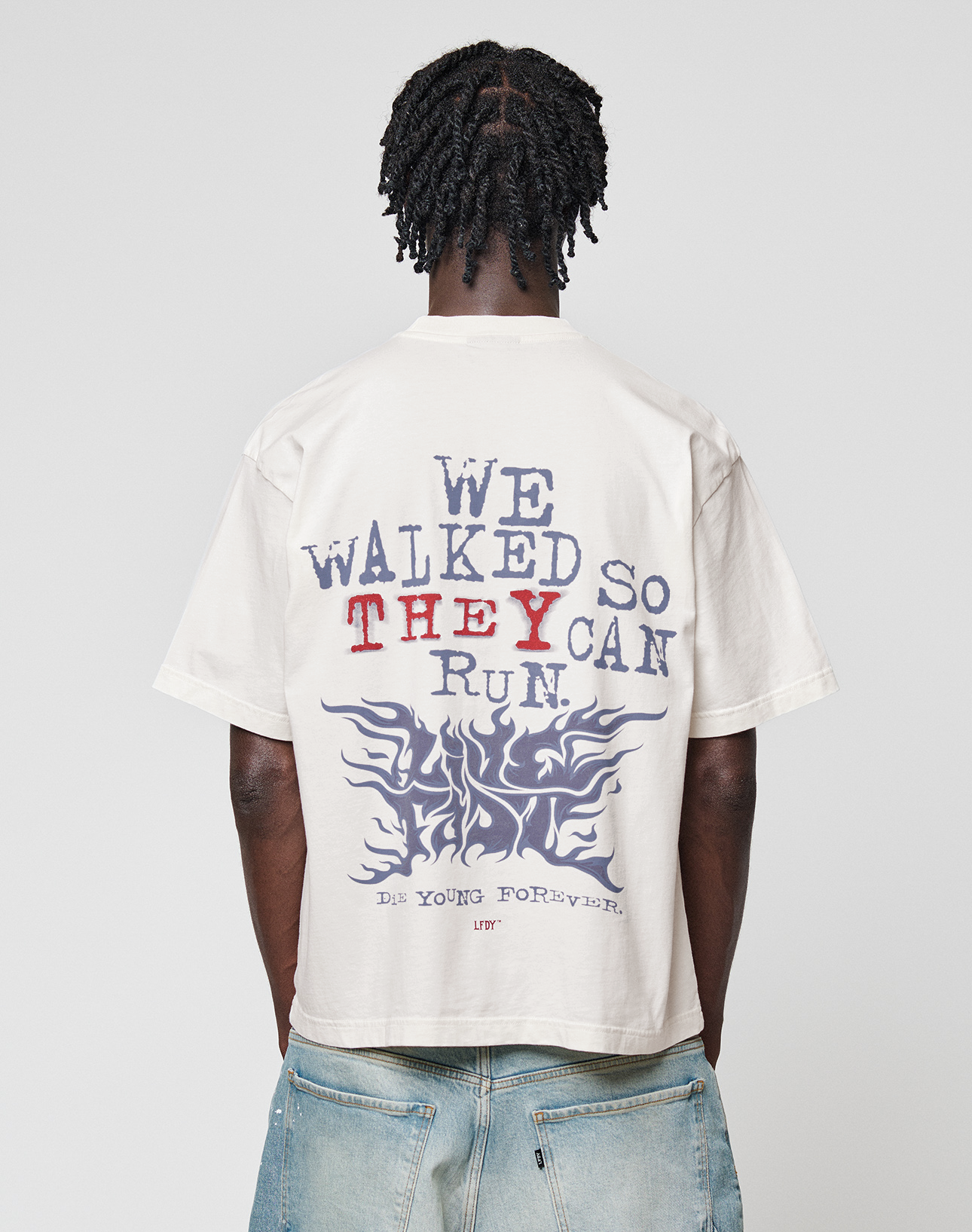 Seen from behind, a person with braided hair wears the LIVE FAST DIE YOUNG LFDY We Walked Tee—an oversize-fit white shirt made of 100% cotton, featuring "WE WALKED SO THEY CAN RUN." and "DIE YOUNG FOREVER" prints with flame graphics—paired with faded jeans. | model