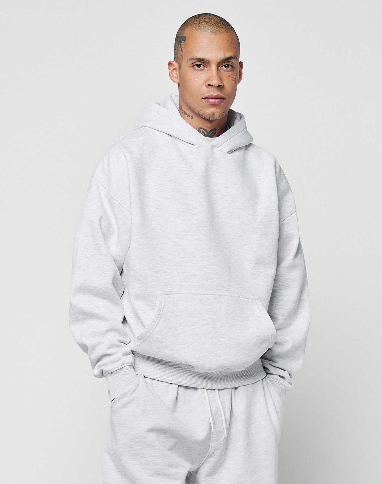 A person with a shaved head and tattoos wears the LIVE FAST DIE YOUNG Basic Blank Hooded sweatshirt and matching pants in light gray, made from a soft cotton-polyester blend, standing against a plain background with hands in the hoodie pocket.