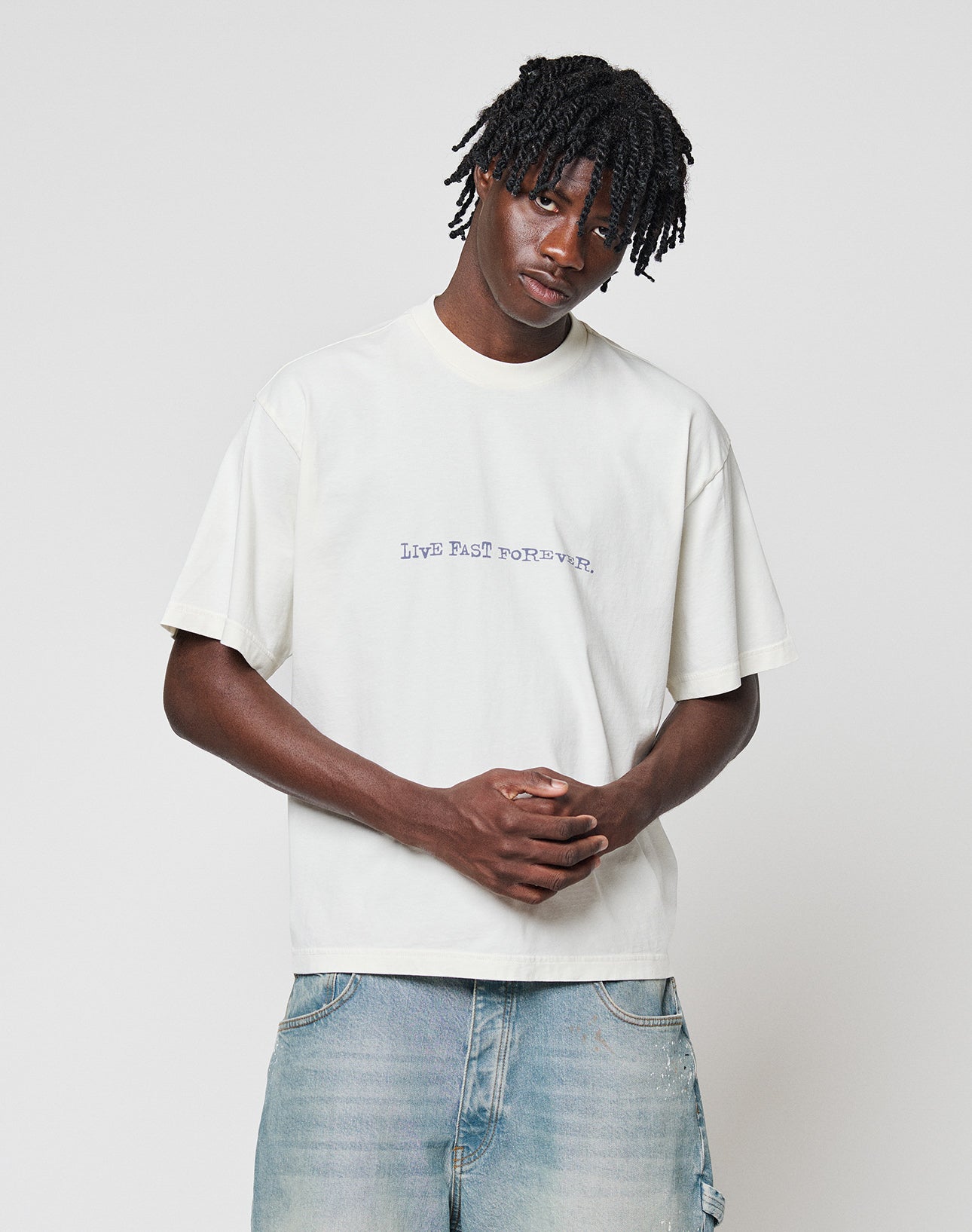 A young man with short dreadlocks wears the LIVE FAST DIE YOUNG LFDY We Walked Tee—an oversized, 100% Baumwolle white shirt with bold front and back LIVE FAST TO RESIST prints—paired with faded blue jeans against a light gray background.