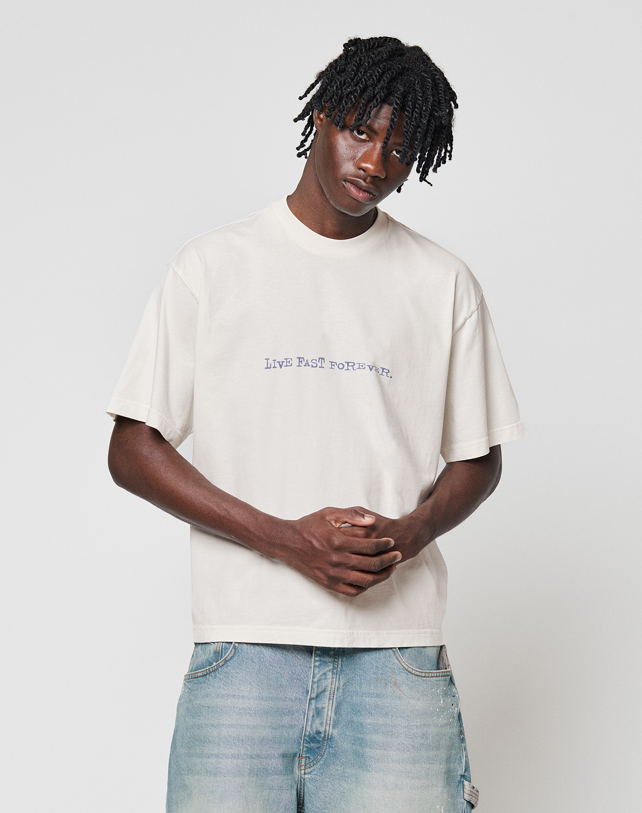 Young man with short dreadlocks wearing an oversize fit, 100% cotton LFDY We Walked Tee by LIVE FAST DIE YOUNG in white with bold front and back print, and light blue jeans, standing against a plain white background with hands clasped.