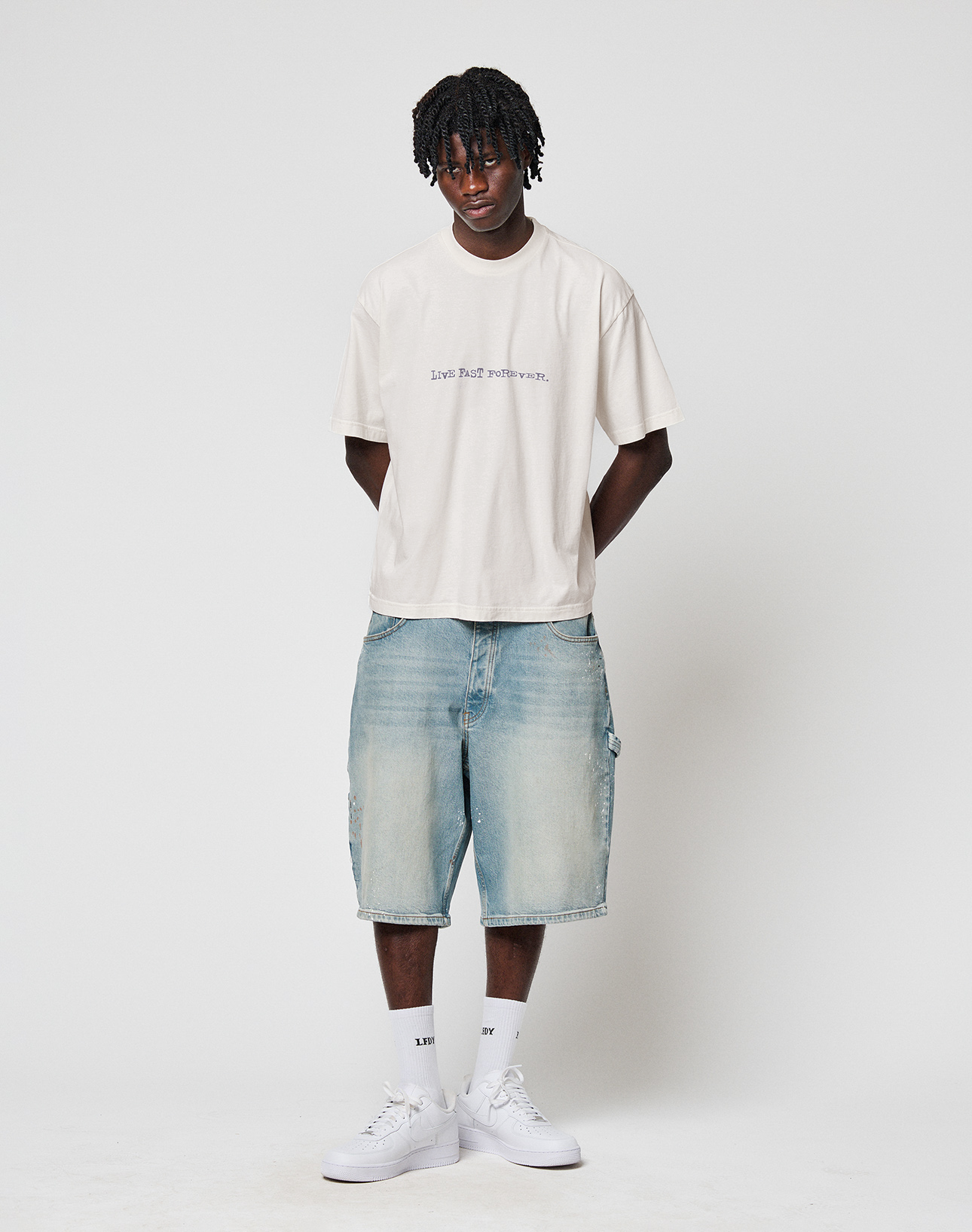A young man wears the LIVE FAST DIE YOUNG LFDY We Walked Tee in an oversize cream fit, paired with loose denim shorts, white socks, and sneakers. He stands against a plain white background with his hands behind his back, looking at the camera.