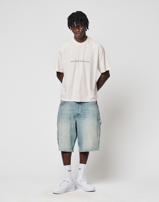 A young man wears the LIVE FAST DIE YOUNG LFDY We Walked Tee in an oversize cream fit, paired with loose denim shorts, white socks, and sneakers. He stands against a plain white background with his hands behind his back, looking at the camera.