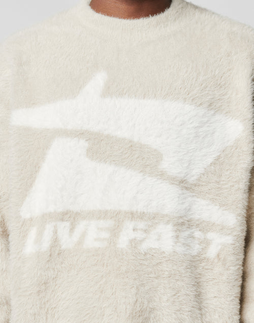 A close-up shows someone in the Live Fast Fuzzy Sweater by LIVE FAST DIE YOUNG, featuring a fuzzy, cream Polyamid-Elastan mix, an oversize fit, and a white intarsia LIVE FAST logo. The persons face isn’t visible.
